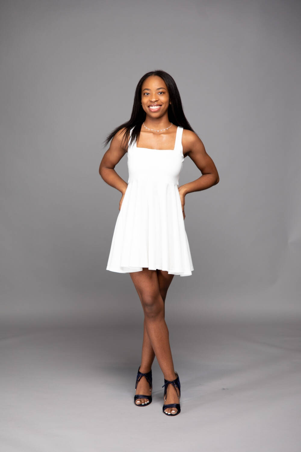 Maria Hall is standing with hands on hips, wearing a white sleeveless dress and black strappy high-heeled sandals, against a plain gray background.